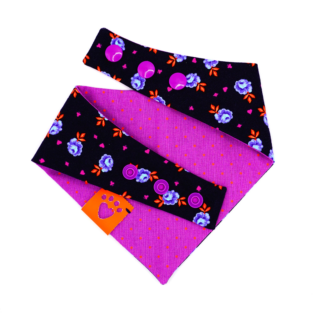 Reversible bandana for dogs. Snaps on back make it adjustable. One side is Navy blue background with purple and light blue rose buds, bright orange leaves and bright purple accent shapes and the other side has Bright purple background with bright orange dots. Orange tag with heart paw cut out on side.
