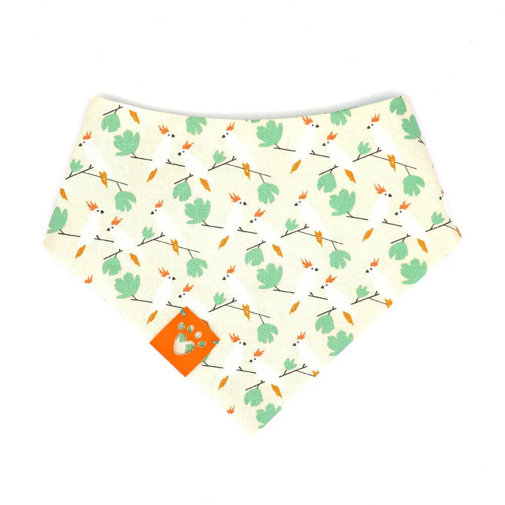 Reversible bandana for dogs. Snaps on back make it adjustable. One side is Light Green background with dark green four leaf clovers and the other side has a Beige background with white, orange and green cockatiel birds. Orange tag with heart paw cut out on side.