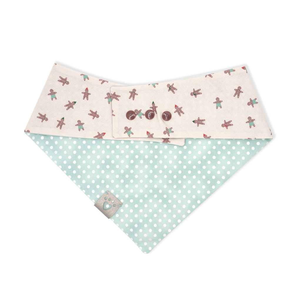 Reversible bandana for dogs. Snaps on back make it adjustable. One side is Distressed minty blue background with white polka dots and the other side has a Very light beige background with brown gingerbread people, some wearing red or black party hats, some with teal pants. Metallic silver tag with heart paw cut out on side.