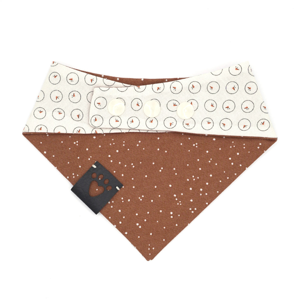 Reversible bandana for dogs. Snaps on back make it adjustable. One side is Light cream background with drawn snowman heads and orange carrot noses and the other side has a Pumpkin orange background with white "snow" dots. Black tag with heart paw cut out on side.