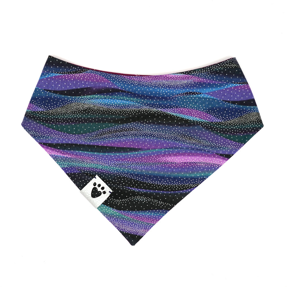 Reversible bandana for dogs. Snaps on back make it adjustable. One side is Black, purple, blue, teal galaxy wave background with silver metallic star dots and the other side has a Purple/wine color background with white constellations and starbursts. Metallic Silver tag with heart paw cut out on side.