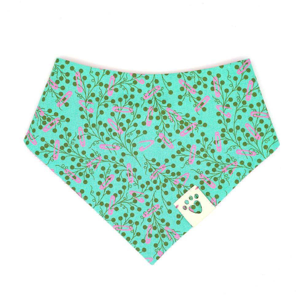 Reversible bandana for dogs. Snaps on back make it adjustable. One side is Teal green background with pink safety pins and dark green flowers and the other side has a Bright pink background with pink, cream and red flowers. Cream tag with heart paw cut out on side.
