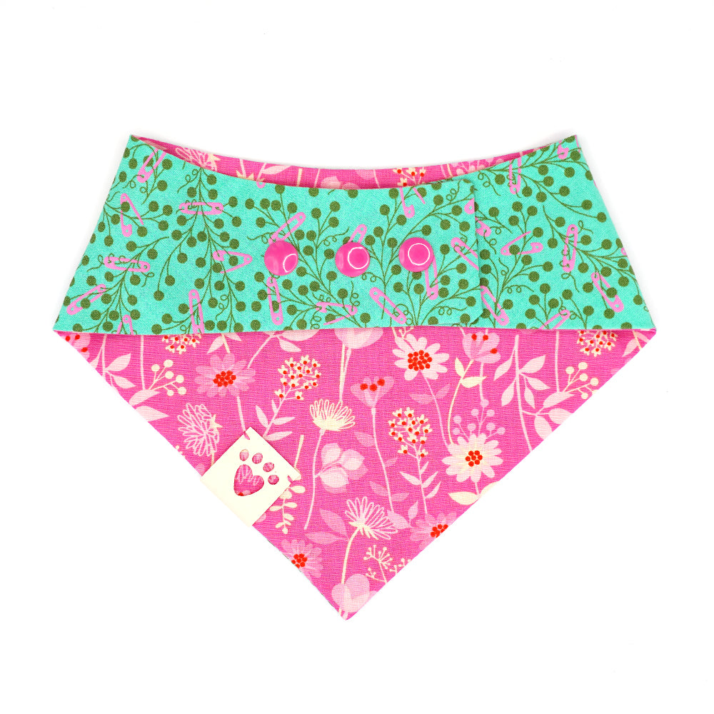 Reversible bandana for dogs. Snaps on back make it adjustable. One side is Teal green background with pink safety pins and dark green flowers and the other side has a Bright pink background with pink, cream and red flowers. Cream tag with heart paw cut out on side.
