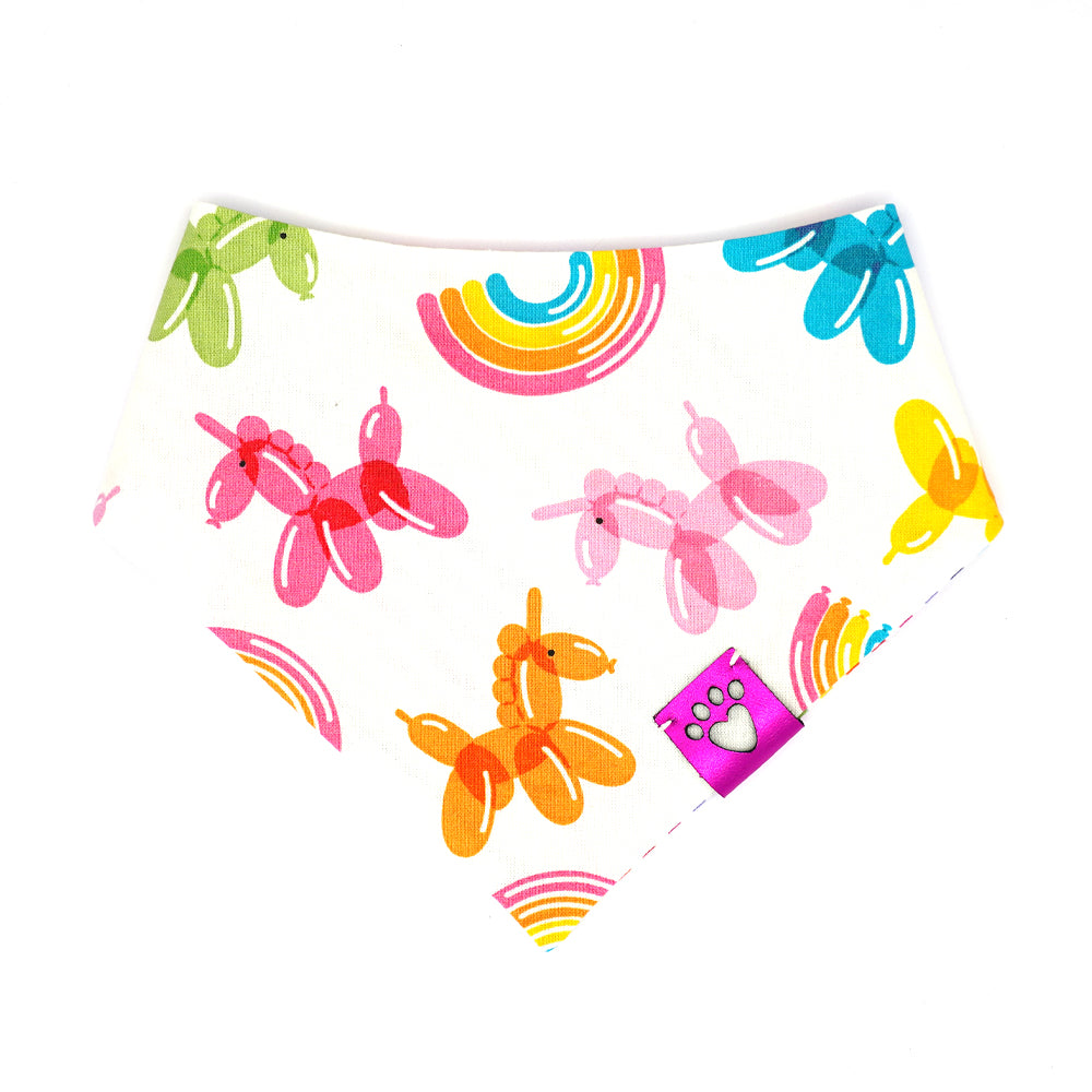 Reversible bandana for dogs. Snaps on back make it adjustable. One side is White background with rainbow stripes and the other side has a White background with rainbow-colored balloon animals and rainbows. Metallic Pink tag with heart paw cut out on side.