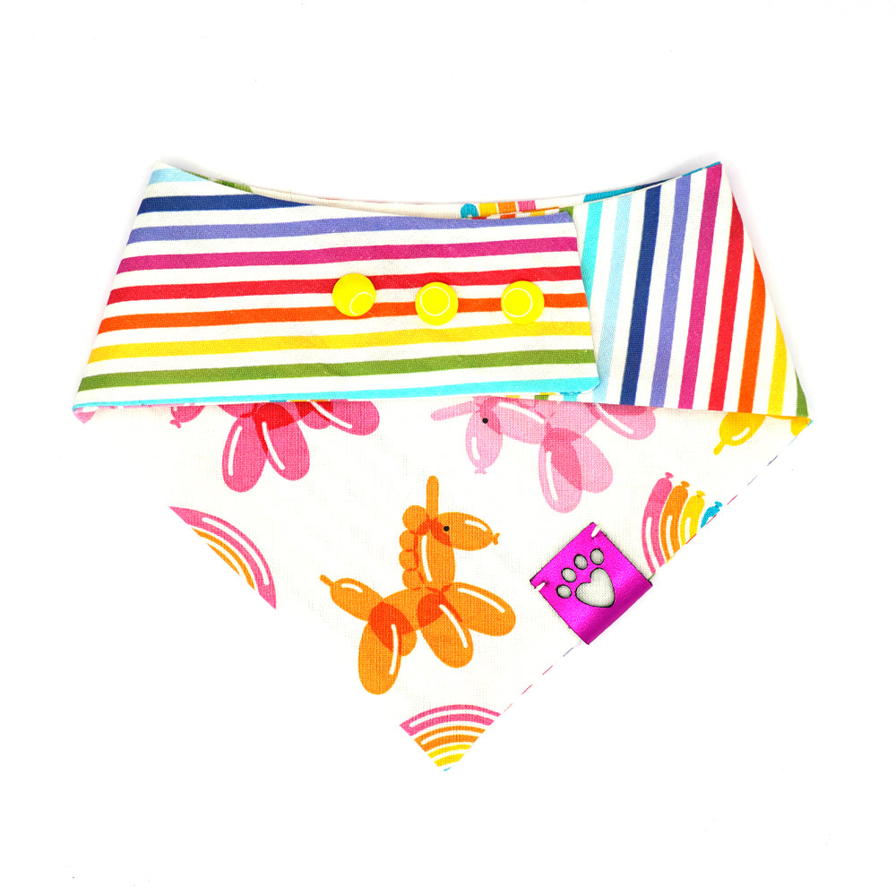 Reversible bandana for dogs. Snaps on back make it adjustable. One side is White background with rainbow stripes and the other side has a White background with rainbow-colored balloon animals and rainbows. Metallic Pink tag with heart paw cut out on side.