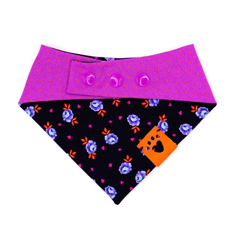 Reversible bandana for dogs. Snaps on back make it adjustable. One side is Navy blue background with purple and light blue rose buds, bright orange leaves and bright purple accent shapes and the other side has Bright purple background with bright orange dots. Orange tag with heart paw cut out on side.