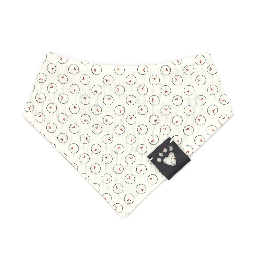 Reversible bandana for dogs. Snaps on back make it adjustable. One side is Light cream background with drawn snowman heads and orange carrot noses and the other side has a Pumpkin orange background with white "snow" dots. Black tag with heart paw cut out on side.