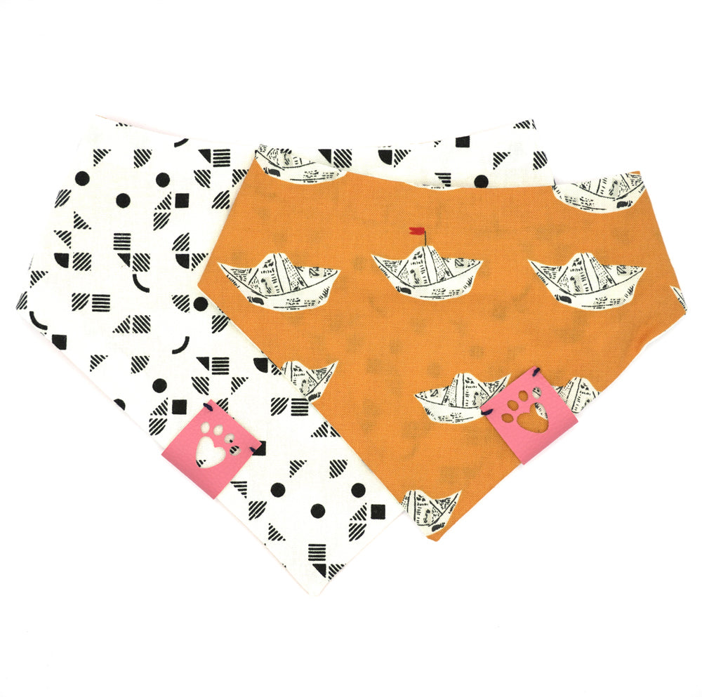 Reversible bandana for dogs. Snaps on back make it adjustable. One side is Bright orange background with newsprint boats and the other side has a White background with black geometric pattern. Light Pink tag with heart paw cut out on side.