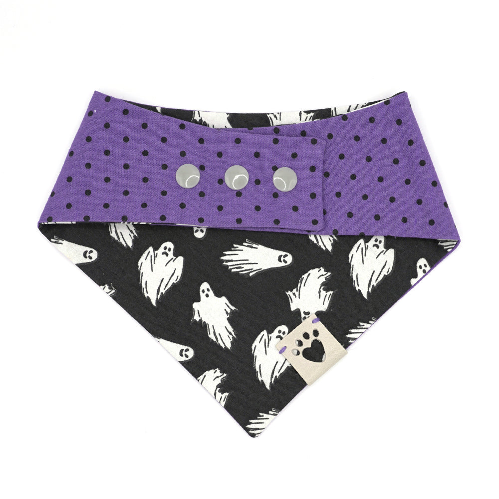 Reversible bandana for dogs. Snaps on back make it adjustable. One side is Black background with white flying ghosts and the other side has a Purple background with black polka dots. Light Grey tag with heart paw cut out on side.