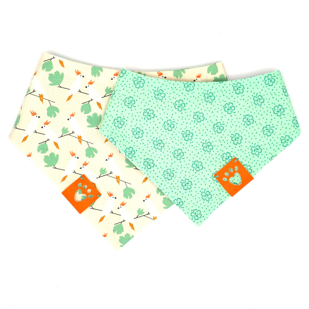 Reversible bandana for dogs. Snaps on back make it adjustable. One side is Light Green background with dark green four leaf clovers and the other side has a Beige background with white, orange and green cockatiel birds. Orange tag with heart paw cut out on side.