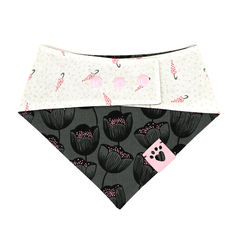 Reversible bandana for dogs. Snaps on back make it adjustable. One side is White background with pink polk-a-dot umbrellas and grey dots and the other side has a Charcoal grey background with black and pink flowers. Light pink tag with heart paw cut out on side.