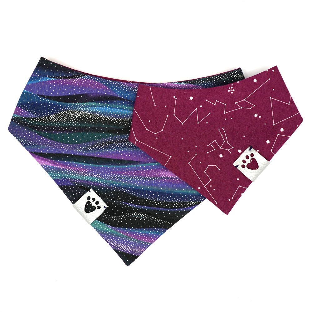 Reversible bandana for dogs. Snaps on back make it adjustable. One side is Black, purple, blue, teal galaxy wave background with silver metallic star dots and the other side has a Purple/wine color background with white constellations and starbursts. Metallic Silver tag with heart paw cut out on side.