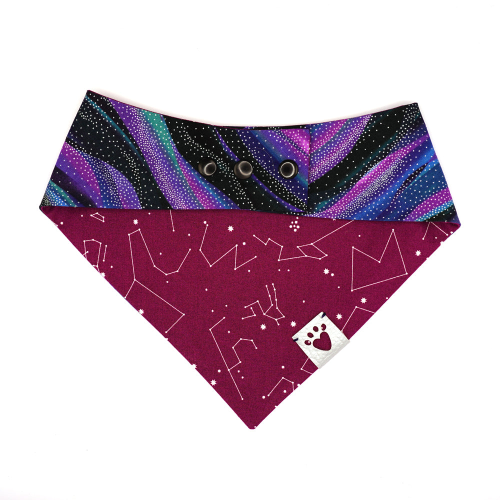 Reversible bandana for dogs. Snaps on back make it adjustable. One side is Black, purple, blue, teal galaxy wave background with silver metallic star dots and the other side has a Purple/wine color background with white constellations and starbursts. Metallic Silver tag with heart paw cut out on side.
