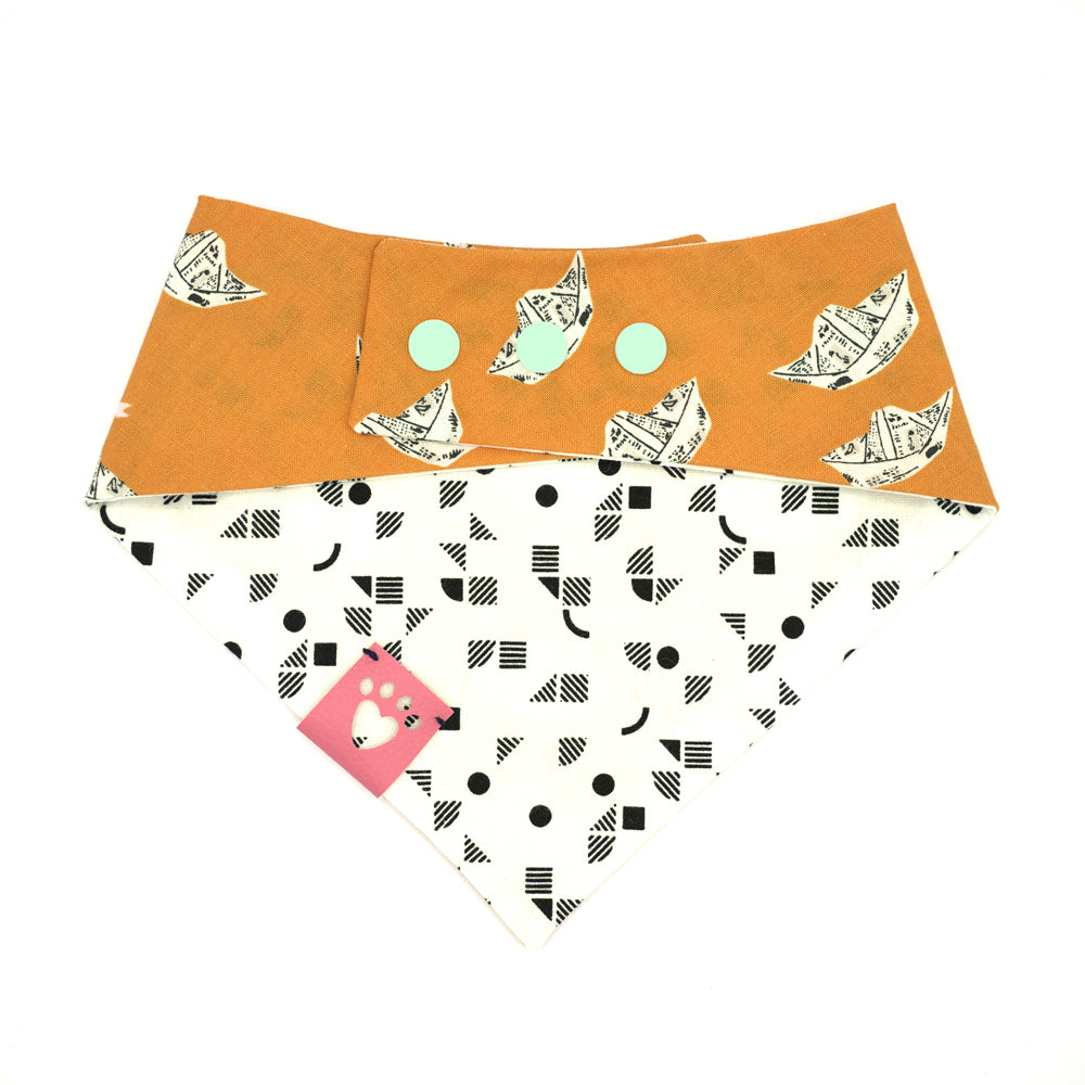 Reversible bandana for dogs. Snaps on back make it adjustable. One side is Bright orange background with newsprint boats and the other side has a White background with black geometric pattern. Light Pink tag with heart paw cut out on side.
