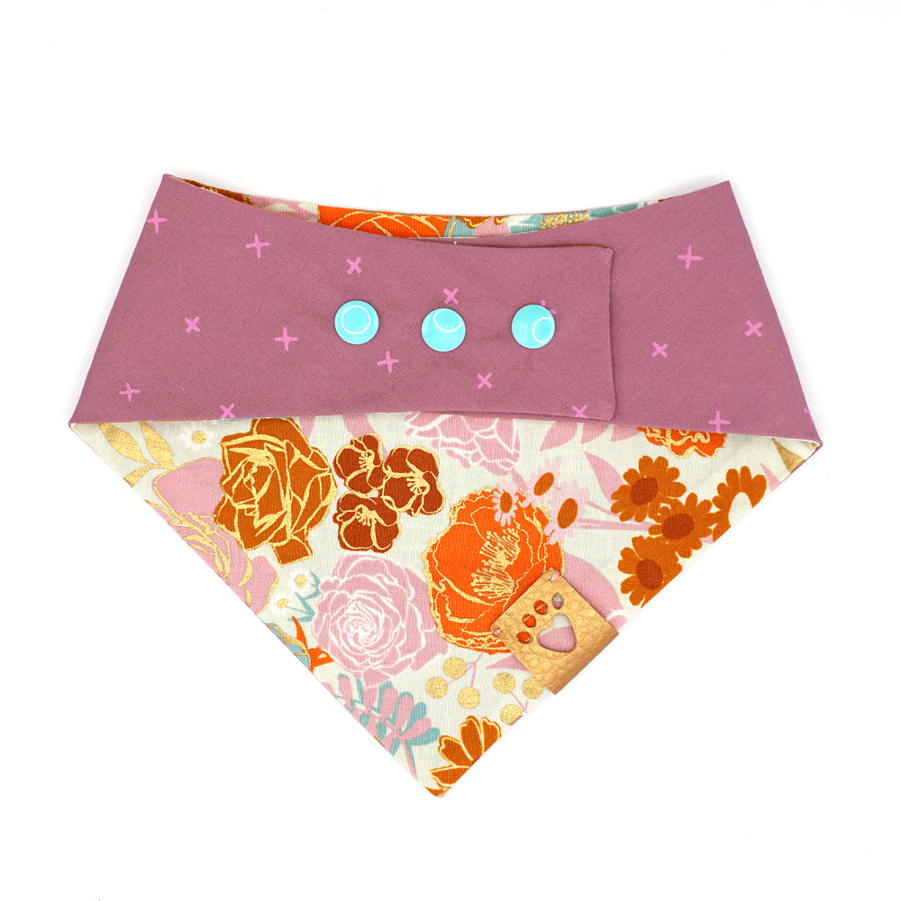 Reversible bandana for dogs. Snaps on back make it adjustable. One side is Light grey background with lilac, teal, pink and orange flowers and gold metallic accents and the other side has a Lilac background with purple/pink Xs. Metallic Gold tag with heart paw cut out on side.