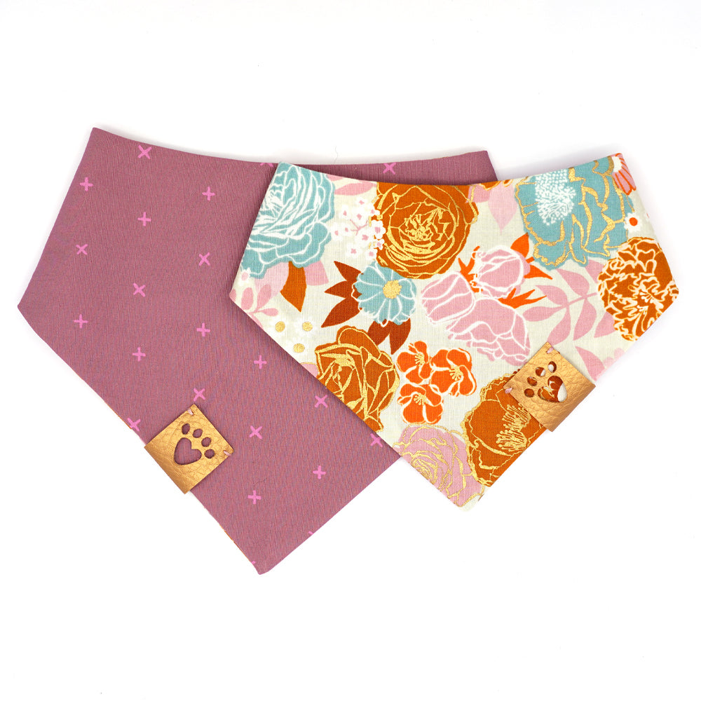 Reversible bandana for dogs. Snaps on back make it adjustable. One side is Light grey background with lilac, teal, pink and orange flowers and gold metallic accents and the other side has a Lilac background with purple/pink Xs. Metallic Gold tag with heart paw cut out on side.