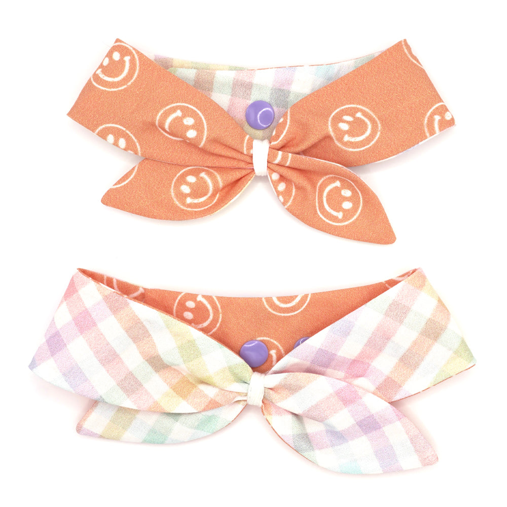 Reversible neckerchief for dogs. Snaps on back make it adjustable. One side has a White background with pastel rainbow plaid pattern and the other side has a Light orange background with White smiley fave illustrations. Embroidery thread wrapped center in white.