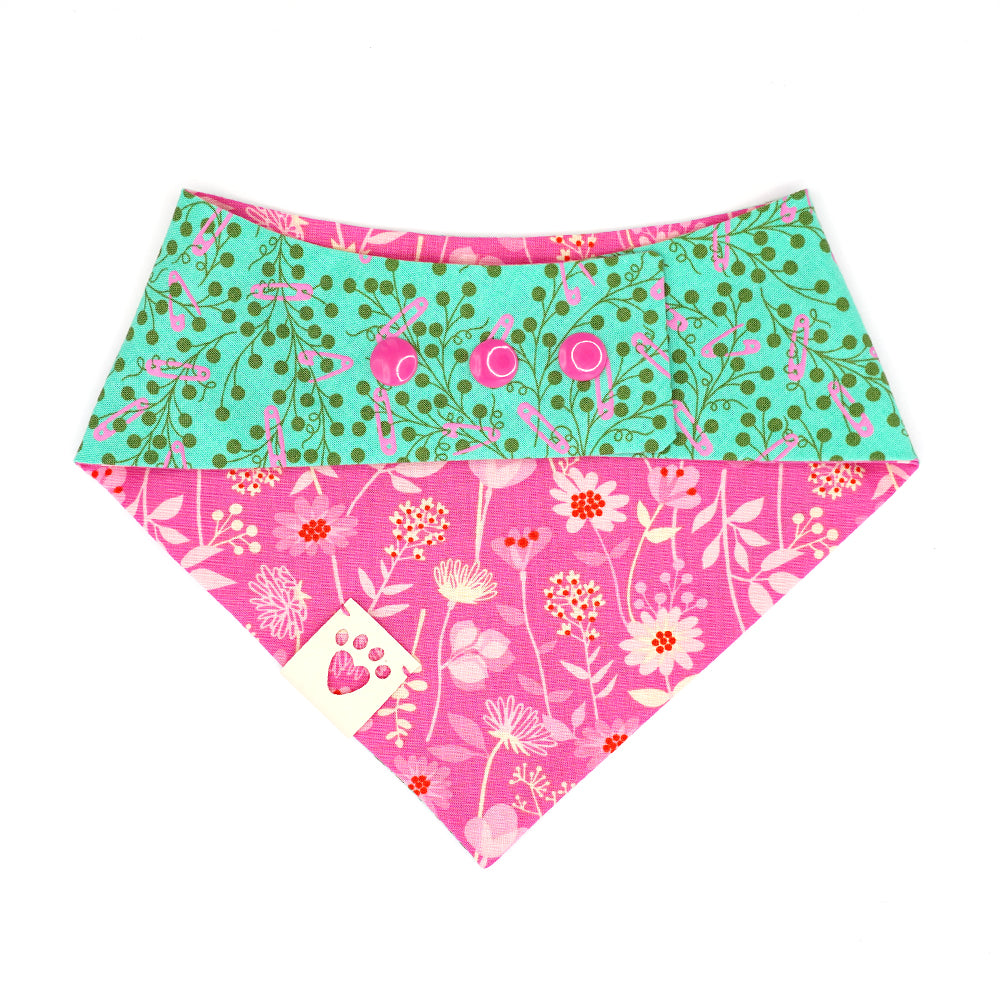  Reversible bandana for dogs. Snaps on back make it adjustable. One side is Teal green background with pink safety pins and dark green flowers and the other side has a Bright pink background with pink, cream and red flowers. Cream tag with heart paw cut out on side.