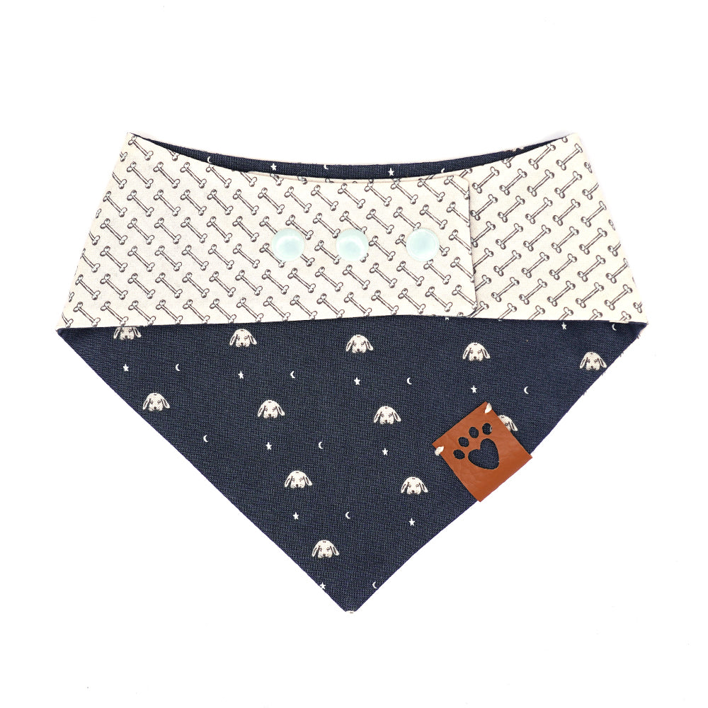 Reversible bandana for dogs. Snaps on back make it adjustable. One side is Navy blue background with tan illustrated dog heads, moons and stars and the other side has a Cream background with black illustrated bones (very pale blue inside bone). Tan tag with heart paw cut out on side.